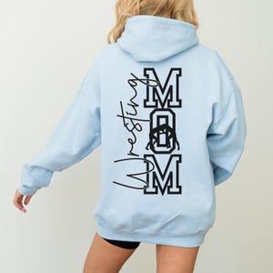 May include: A light blue hooded sweatshirt with the words "Wrestling Mom" printed on the back in black. The "M" in Mom is stylized with a wrestler silhouette.
