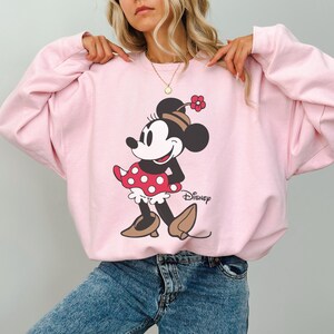 May include: A pale pink sweatshirt with a cartoon image of Minnie Mouse. Minnie is wearing a red polka dot dress, brown shoes, and a hat with a flower. The Disney logo is printed in black on the bottom right.