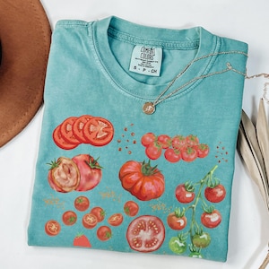 May include: A teal blue t-shirt with a tomato graphic. The shirt has a distressed look and features a variety of tomato shapes and sizes, including whole tomatoes, tomato slices, and cherry tomatoes. The shirt also has the text "COMFORT COLORS" and "S - P - CH" printed on the tag.