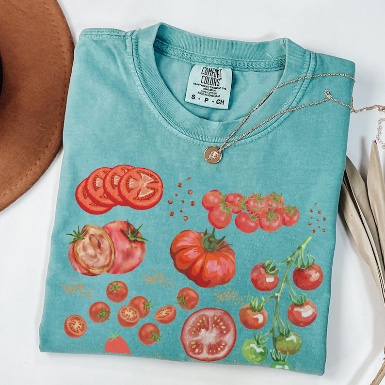 May include: A teal blue t-shirt with a tomato graphic. The shirt has a distressed look and features a variety of tomato shapes and sizes, including whole tomatoes, tomato slices, and cherry tomatoes. The shirt also has the text "COMFORT COLORS" and "S - P - CH" printed on the tag.