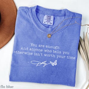 May include: A periwinkle blue short-sleeved t-shirt featuring the white printed text: You are enough. And anyone who tells you otherwise isn't worth your time, followed by a signature and a butterfly graphic. A gold-toned necklace rests on the shirt.