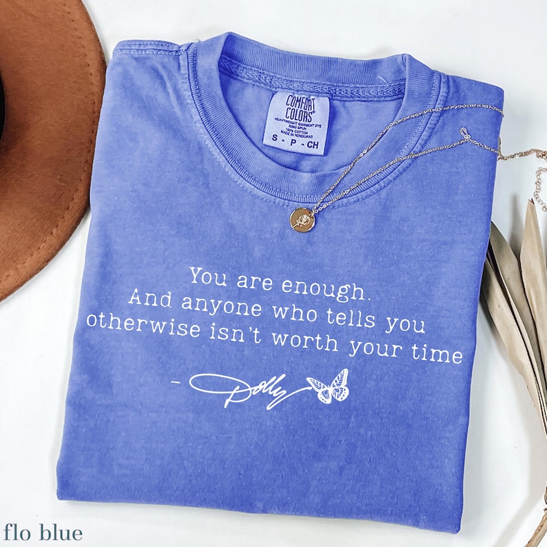 May include: A periwinkle blue short-sleeved t-shirt featuring the white printed text: You are enough. And anyone who tells you otherwise isn't worth your time, followed by a signature and a butterfly graphic. A gold-toned necklace rests on the shirt.