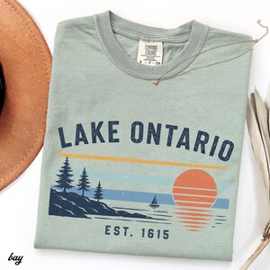 May include: A sage green short-sleeve t-shirt featuring the text LAKE ONTARIO and EST. 1615. The graphic includes a dark blue shoreline with pine trees, a small sailboat, and a large orange sun with horizontal lines reflecting on the water.