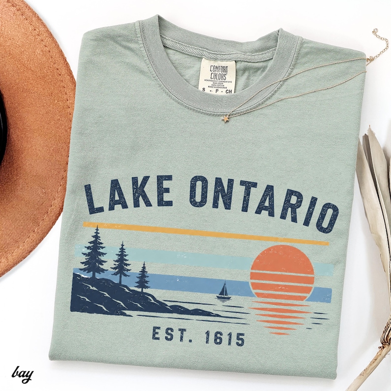 May include: A sage green short-sleeve t-shirt featuring the text LAKE ONTARIO and EST. 1615. The graphic includes a dark blue shoreline with pine trees, a small sailboat, and a large orange sun with horizontal lines reflecting on the water.
