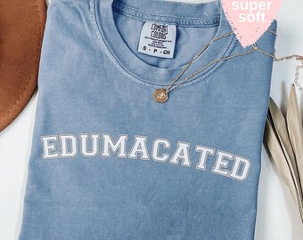 May include: A light blue Comfort Colors t-shirt with the word "EDUMACATED" in white block letters. A gold necklace with a pendant rests on the shirt. The shirt is labeled "super soft" with a pink heart-shaped sticker.