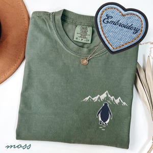 May include: A moss green t-shirt with an embroidered penguin and mountain design. The shirt has a crew neck and short sleeves. A denim heart-shaped patch with the word "Embroidery" is visible. A gold necklace with a pendant is also present.