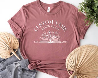 May include: A dusky pink t-shirt featuring the text "CUSTOM NAME BOOK CLUB" above a floral book design. The shirt includes the year "2025" and is styled with jeans and decorative fans.