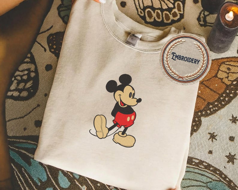 May include: A beige sweatshirt with an embroidered Mickey Mouse design. The cartoon character is depicted in his iconic outfit, with black ears, red shorts, and yellow shoes. A circular patch with the word "EMBROIDERY" is also visible.