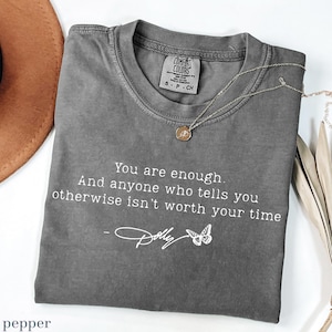 May include: A charcoal grey short-sleeved t-shirt featuring the white printed text: You are enough. And anyone who tells you otherwise isn't worth your time - Dolly, accompanied by a small butterfly graphic. The shirt has a crew neckline.