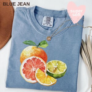 Op de afbeelding: Een lichtblauw T-shirt met een aquarel illustratie van citrusvruchten, waaronder een sinaasappel, grapefruit en limoen. Het shirt heeft de tekst "BLUE JEAN" linksboven en een gouden ketting. Het label luidt "COMFORT COLORS".
