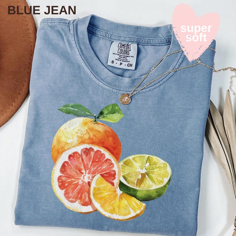 Op de afbeelding: Een lichtblauw T-shirt met een aquarel illustratie van citrusvruchten, waaronder een sinaasappel, grapefruit en limoen. Het shirt heeft de tekst "BLUE JEAN" linksboven en een gouden ketting. Het label luidt "COMFORT COLORS".