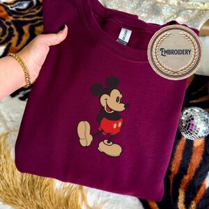 May include: A maroon sweatshirt with an embroidered Mickey Mouse design. The cartoon character is depicted in his iconic outfit. The sweatshirt has a crew neck and a "Gildan" tag. The word "Embroidery" is on a circular patch. The image shows Mickey Mouse walking with a smile.