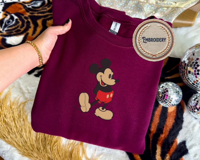 May include: A maroon sweatshirt with an embroidered Mickey Mouse design. The cartoon character is depicted in his iconic outfit. The sweatshirt has a crew neck and a "Gildan" tag. The word "Embroidery" is on a circular patch. The image shows Mickey Mouse walking with a smile.