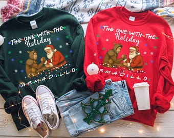 May include: Two long-sleeved sweatshirts, one green and one red, with a holiday-themed graphic. The green sweatshirt has the text "The One With The Holiday Armadillo" and a Santa Claus and armadillo design. The red sweatshirt has the same design and text. White sneakers and blue jeans are also visible.