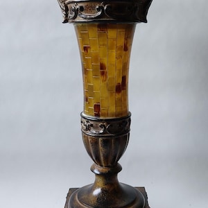 Può includere: Un piedistallo ornato color bronzo, con base quadrata e sommità scanalata a forma di urna. La colonna centrale presenta un mosaico di piastrelle gialle e marroni. Elementi decorativi: volute e bordo dettagliato.