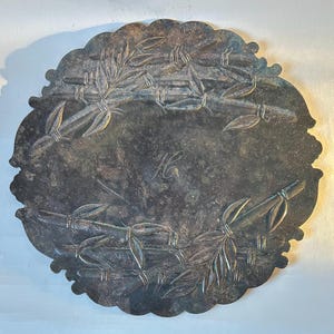 May include: Silver-toned, oval-shaped tray with a scalloped edge. The tray features an embossed bamboo design and a central monogram. The surface shows signs of age and wear, adding to its vintage appeal.