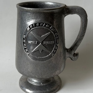 Peut inclure: Une chope en métal de couleur étain avec une anse et une base en piédestal. La chope présente un emblème en relief avec le texte "Material ID Company Metal Division" et un motif d'étoile stylisé. La chope a un aspect classique et vintage.