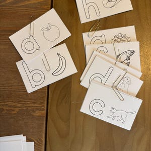 May include: White flashcards with black outline illustrations of an apple, banana, cowboy hat, gift, fish, dog, cat, and letters a, b, c, d, h. The cards are held together with paper clips and are arranged on a wooden surface.