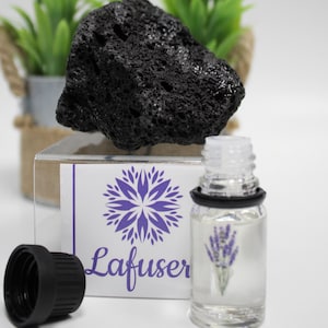 Puede incluir: Un difusor de roca de lava negra con una pequeña botella de aceite esencial de lavanda. La botella tiene una tapa negra y una etiqueta con un diseño de flor morada y el texto "Lafuser".