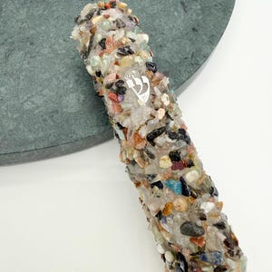 May include: A cylindrical object covered in colorful, small stones. The stones are various colors, including red, blue, and brown. A silver emblem is visible on the side. The object is resting on a green, circular surface.