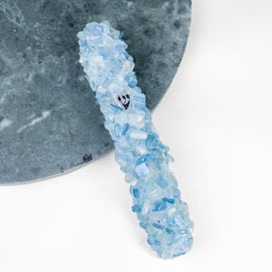 Może przedstawiać: Jasnoniebieska kasetka na mezuzę pokryta małymi, nieregularnymi niebieskimi i przezroczystymi kamieniami. Mały, czarno-biały emblemat jest widoczny z przodu. Mezuza spoczywa na szarej, okrągłej powierzchni.