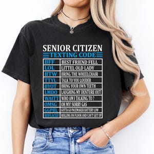 May include: Black t-shirt with the text "SENIOR CITIZEN TEXTING CODE" in white and blue. The shirt lists texting acronyms and their meanings, such as "BFF" for "BEST FRIEND FELL" and "LOL" for "LITTEL OLD LADY".