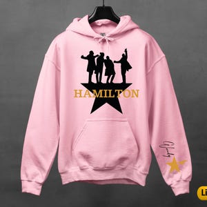 A pink hoodie with a graphic design featuring silhouettes of three people and the word "HAMILTON" printed on it.