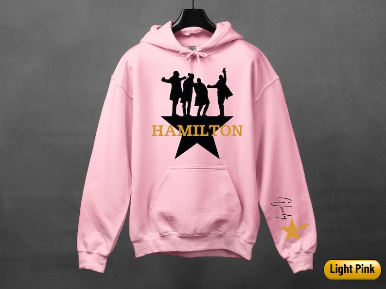 A pink hoodie with a graphic design featuring silhouettes of three people and the word "HAMILTON" printed on it.