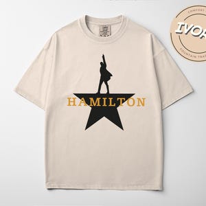 A beige t-shirt with a graphic design featuring a silhouette of a person standing on a star and the text "HAMILTON" below it.