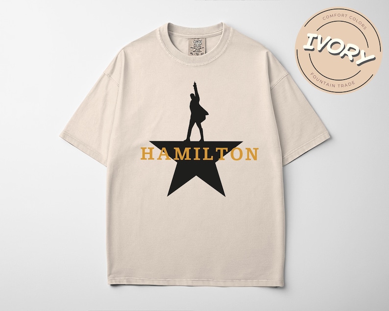 A beige t-shirt with a graphic design featuring a silhouette of a person standing on a star and the text "HAMILTON" below it.