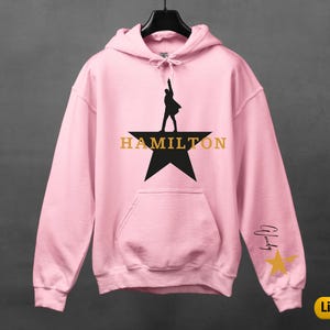 A pink hoodie with a black silhouette of a person holding a microphone and the word "HAMILTON" printed on it, along with a star and a yellow star.