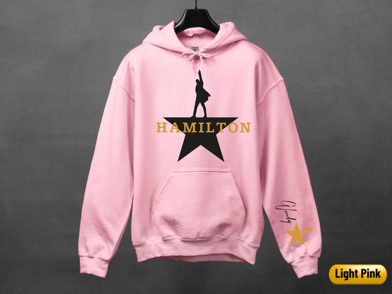 A pink hoodie with a black silhouette of a person holding a microphone and the word "HAMILTON" printed on it, along with a star and a yellow star.