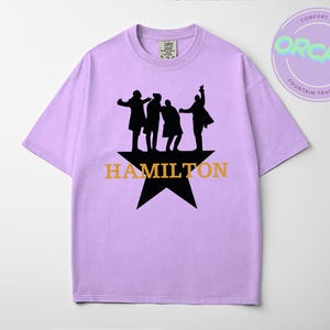 A purple t-shirt with a graphic design featuring silhouettes of four people and the text "HAMILTON" in orange letters.