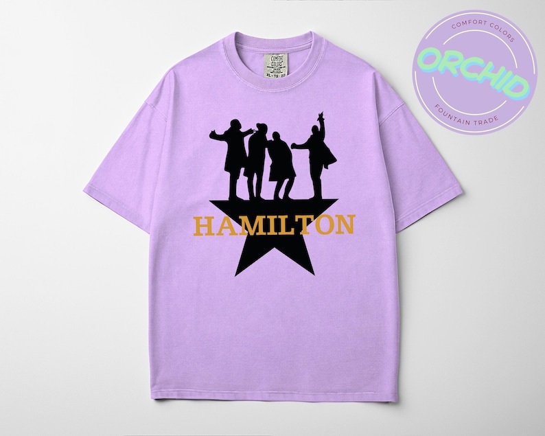 A purple t-shirt with a graphic design featuring silhouettes of four people and the text "HAMILTON" in orange letters.