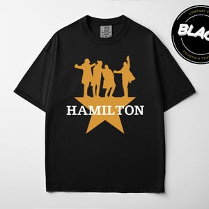 A black t-shirt with a graphic design featuring silhouettes of three people and a star, along with the text "HAMILTON" printed on it.