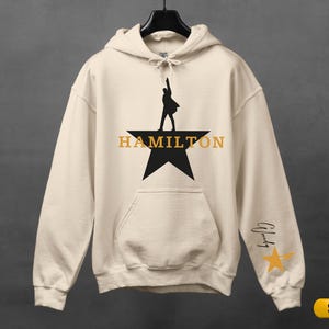 A white hoodie with a black silhouette of a person on a star and the word "HAMILTON" printed on it.