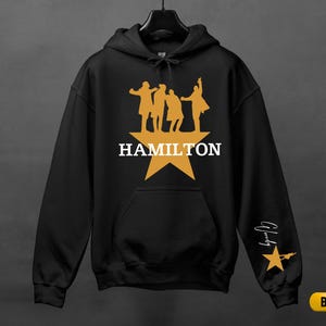 A black hoodie with a yellow star and silhouettes of three people, along with the text "HAMILTON" and a star.