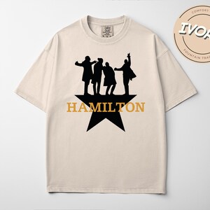 A beige t-shirt with a graphic design featuring silhouettes of three people and the text "HAMILTON" in orange letters.