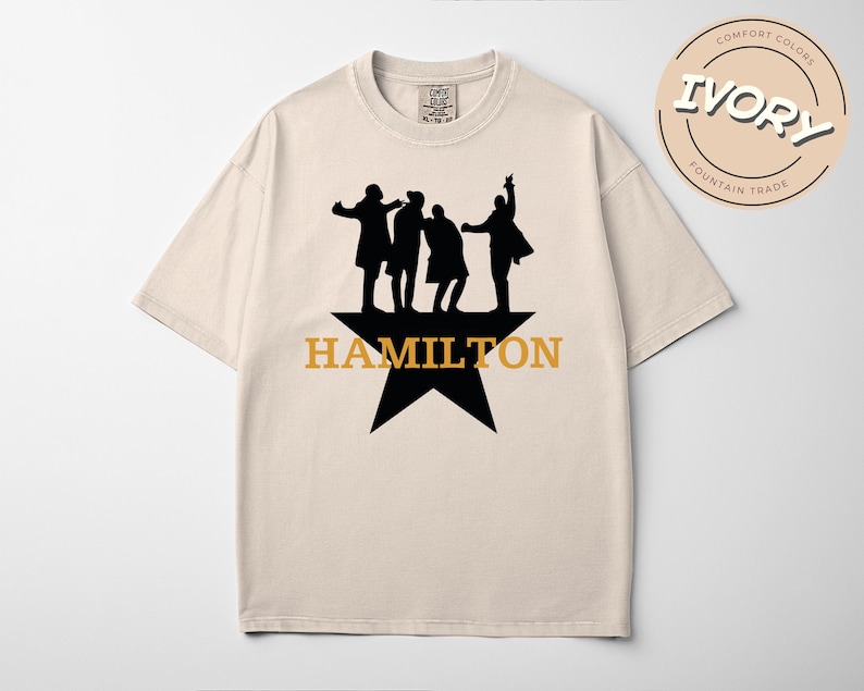A beige t-shirt with a graphic design featuring silhouettes of three people and the text "HAMILTON" in orange letters.