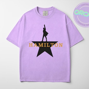 A purple t-shirt with a graphic design featuring a silhouette of a person standing on a star and the word "HAMILTON" written below.