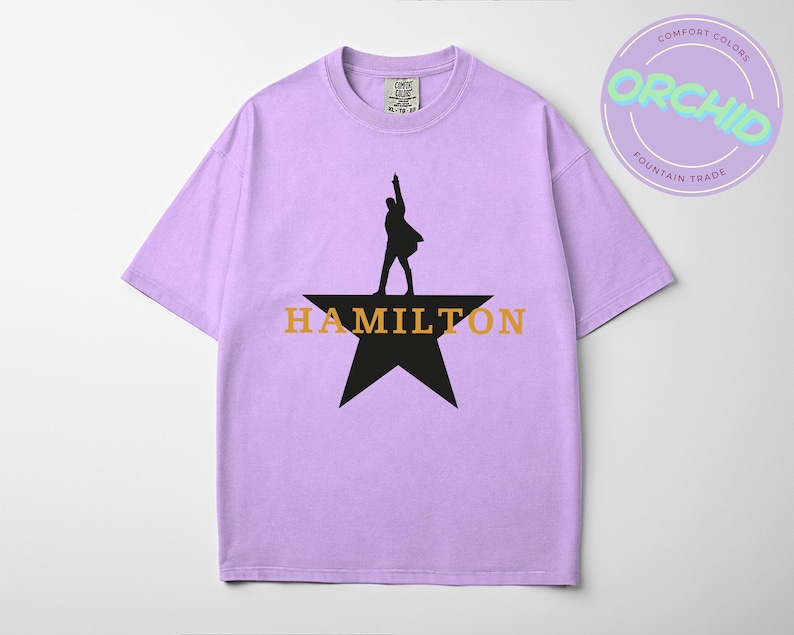 A purple t-shirt with a graphic design featuring a silhouette of a person standing on a star and the word "HAMILTON" written below.