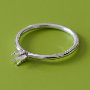 May include: A silver ring with a prong setting, designed to hold a gemstone. The ring is set against a solid green background. The ring's band is smooth and circular, and the setting has four prongs.