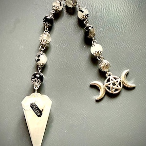 May include: A pendulum featuring a clear, faceted crystal point with a black inclusion, attached to a chain of alternating black and white beads with silver-toned metal caps. The chain ends with a silver-toned triple moon and pentagram charm.