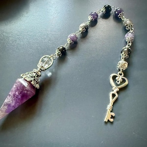 May include: A pendulum featuring a pointed purple amethyst crystal attached to a silver-toned metal chain. The chain is adorned with alternating dark and light purple beads and a silver-toned metal key charm with a heart-shaped top.