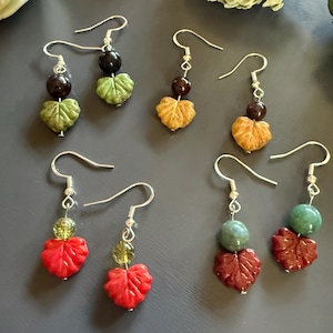 May include: A collection of handcrafted earrings featuring leaf and bead designs. The earrings showcase a variety of colors, including green, red, and orange, with silver-tone ear wires. The leaf-shaped elements are intricately carved, adding a unique touch to each pair.
