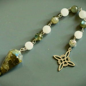 May include: A pendulum featuring a faceted labradorite point, alternating white and labradorite beads, and a silver-toned metal charm with a four-pointed knot design. The components are connected by silver-toned metal links and wire loops.