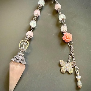 May include: A pendulum featuring a pointed rose quartz crystal capped with ornate silver-toned metal. The chain consists of alternating pale pink and translucent white beads with silver caps, a coral-colored rose charm, a silver butterfly, and a small charm.