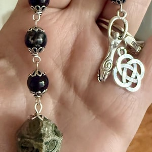 May include: A pendulum featuring a faceted, mottled green and brown stone point suspended from a silver-toned chain. The chain includes dark purple spherical beads encased in ornate silver-toned caps, a silver-toned goddess charm, and a silver-toned Celtic knot charm.
