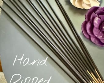 May include: Hand-dipped incense sticks in a bundle, with gold-colored tips, arranged on a gray surface. The image includes decorative, handcrafted flower accents in shades of pink, green, cream, and purple.