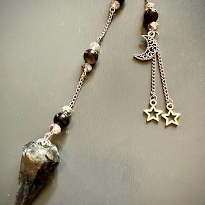 May include: A pendulum featuring a faceted, dark gray and black stone point suspended from a silver-toned metal chain. The chain is accented with round black beads, smaller clear beads, and a silver-toned crescent moon charm with two dangling star charms.